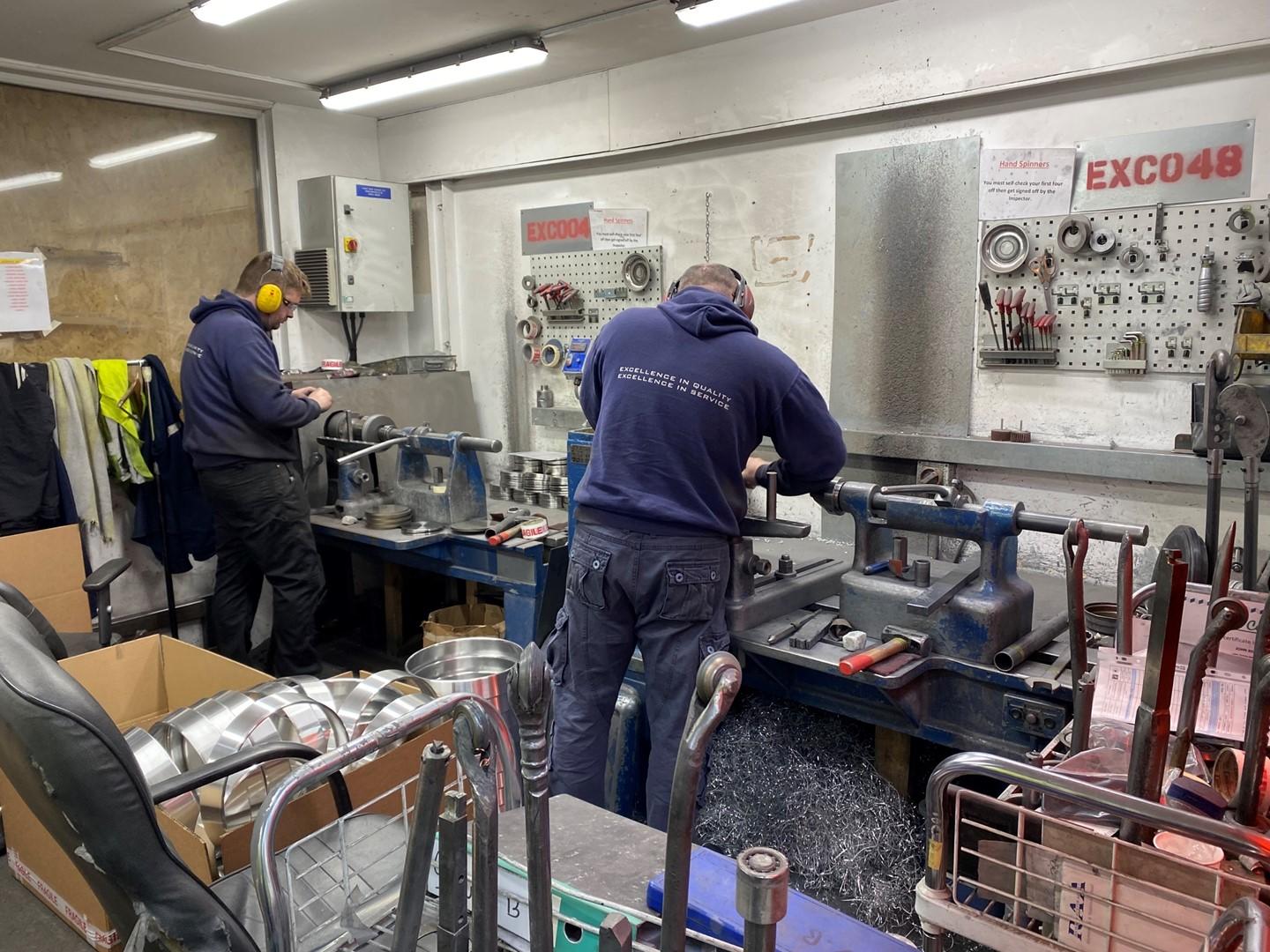 Two men in blue hooded sweaters and ear protective wear working in a metal spinning workshop